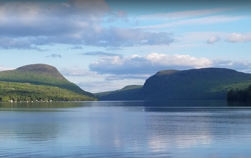A serene lake surrounded by green hills and mountains under a blue sky with scattered clouds.