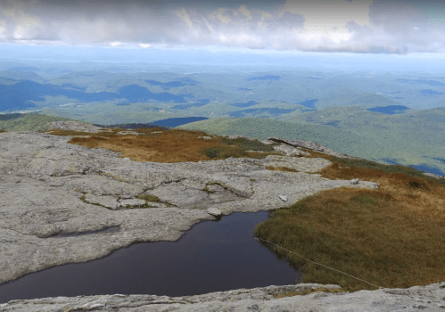 A panoramic view of rolling green mountains under a cloudy sky, with a rocky foreground and a small pond.