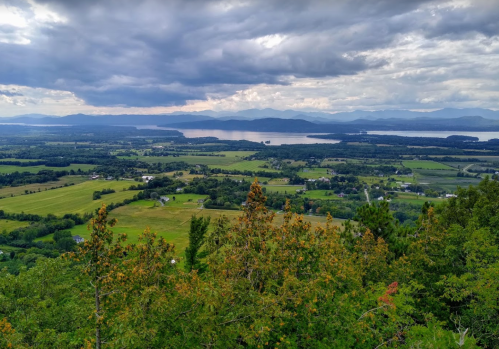 A panoramic view of lush green fields, a river, and distant mountains under a cloudy sky.
