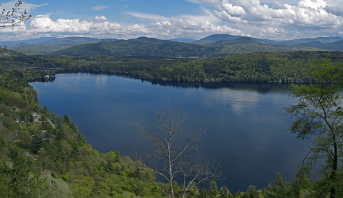 A panoramic view of a serene lake surrounded by lush green hills and mountains under a partly cloudy sky.