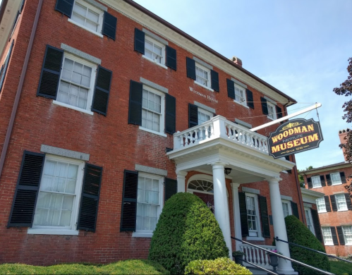Historic red brick building with black shutters and a sign reading "Woodman Museum" hanging from a porch.