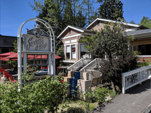 A charming restaurant with outdoor seating, surrounded by greenery and a clear blue sky.