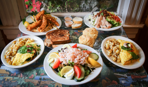 A spread of various dishes including fried fish, salads, omelets, and sides on a table by a window.