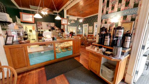 Cozy café interior with wooden furniture, a display case of baked goods, and a coffee station. Floral wallpaper adds charm.