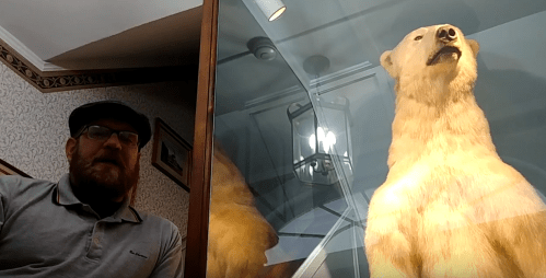 A man sits beside a mounted polar bear in a display case, with a vintage interior in the background.