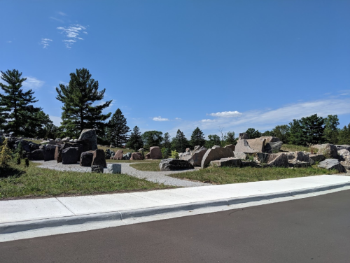 A rocky landscape with large boulders and trees under a clear blue sky, next to a paved road.