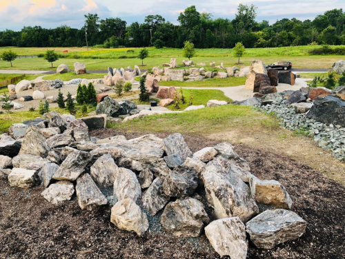 A scenic view of a landscaped area featuring large rocks, greenery, and a gravel path under a cloudy sky.