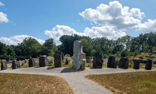 A circle of large stones stands in a grassy area under a blue sky with fluffy clouds and trees in the background.