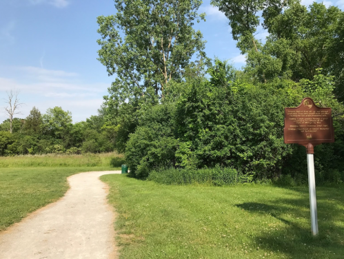 A gravel path leads through green grass and trees, with a historical marker on the right under a clear blue sky.