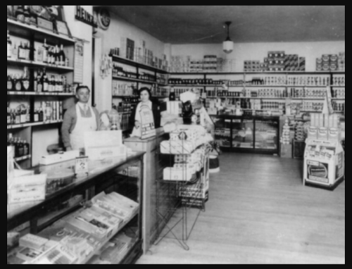 A vintage grocery store interior with shelves full of canned goods and three employees standing behind the counter.