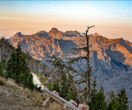 A scenic mountain landscape at sunset, featuring rugged peaks and a winding dirt path surrounded by trees.