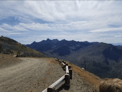 A scenic mountain view with a dirt path and wooden fence under a blue sky with wispy clouds.