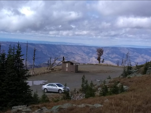 A silver car parked near a small building on a mountain overlook, with distant hills and cloudy skies in the background.