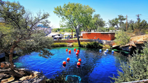 A serene blue water body surrounded by trees, with colorful buoys floating and a gathering area in the background.