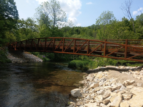 A rust-colored bridge spans a calm river, surrounded by lush green trees and rocky riverbanks under a blue sky.