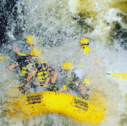 A group of people in yellow helmets white-water rafting, splashing through turbulent water.