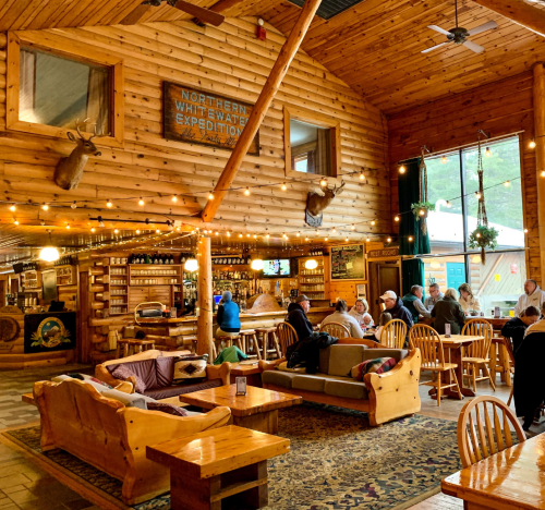 Cozy wooden lodge interior with rustic decor, seating areas, and patrons enjoying food and drinks. Warm, inviting atmosphere.