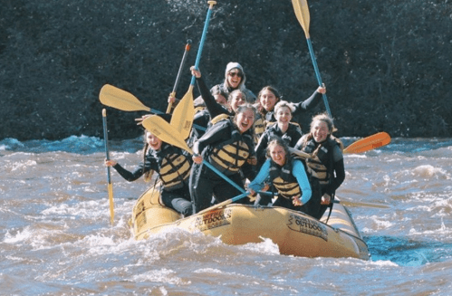 A group of women joyfully rafting on a river, paddling together in a yellow raft amidst splashing water.