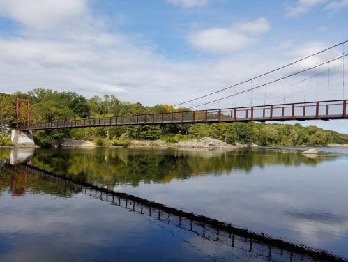 A red suspension bridge spans a calm river, surrounded by trees and reflecting in the water under a cloudy sky.