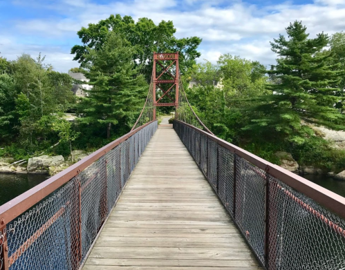 A wooden suspension bridge with a red metal tower, surrounded by trees and a calm river under a partly cloudy sky.
