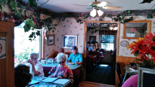 A cozy restaurant interior with diners at tables, decorated with plants and floral arrangements.