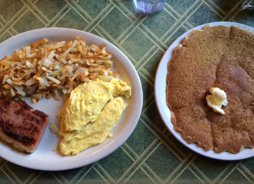 A plate of breakfast featuring scrambled eggs, hash browns, a slice of ham, and a large pancake with butter.