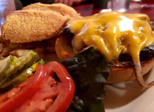 A close-up of a cheeseburger with melted cheese, lettuce, tomato, pickles, and a toasted bun, served with a side of bread.