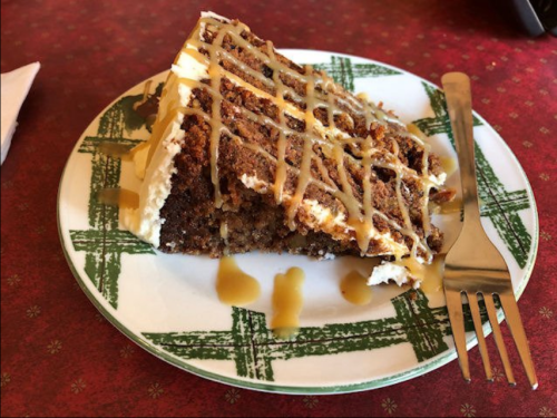 A slice of cake with cream frosting and caramel drizzle on a decorative plate, with a fork beside it.
