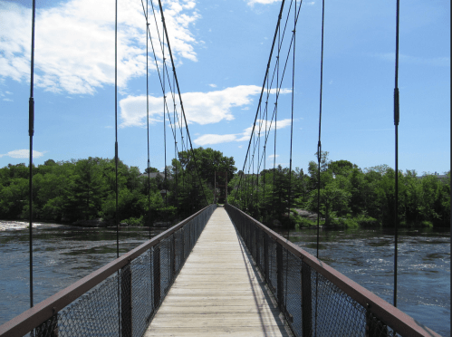 A wooden suspension bridge stretches over a river, surrounded by lush green trees and a clear blue sky.