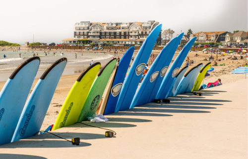 A row of colorful surfboards lined up on the beach, with people enjoying the water and a hotel in the background.