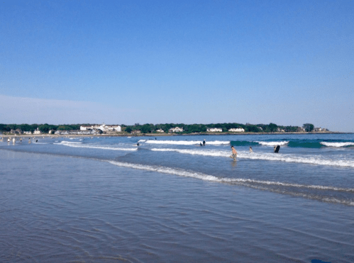 A sunny beach scene with gentle waves, people swimming, and a clear blue sky in the background.