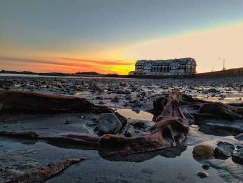 A rocky shoreline at sunset, with a building in the background and colorful sky reflecting on the water.