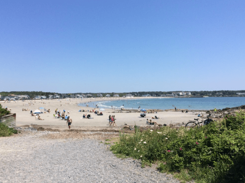 A sunny beach scene with people sunbathing, swimming, and enjoying the shoreline under a clear blue sky.