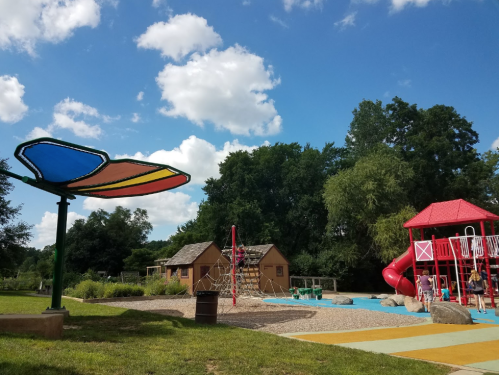 A colorful playground with a slide, climbing structure, and a shaded area, surrounded by trees and blue skies.