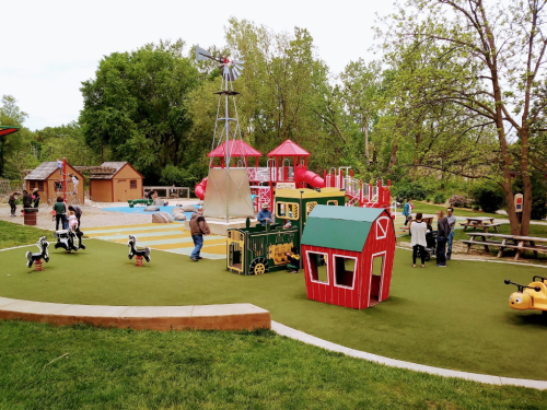A colorful playground with a train, barn, and windmill, surrounded by trees and families enjoying outdoor activities.