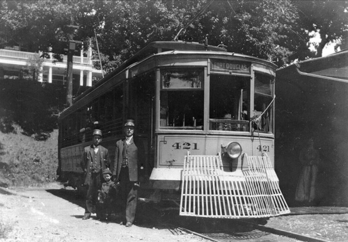 Two conductors and a child stand beside an early 20th-century streetcar labeled "421" in a historic setting.