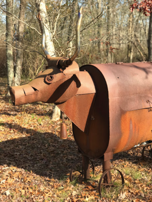 A rusty metal sculpture of a cow with horns, set in a wooded area surrounded by fallen leaves.