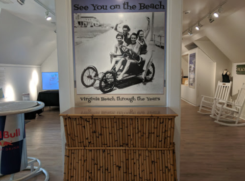 A vintage photo of four people on a beach bike, with a sign reading "See You on the Beach" in a museum setting.