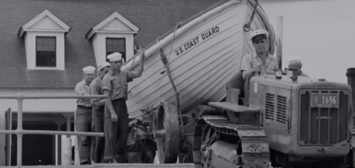 Historical black-and-white image of men lifting a boat onto a tractor near a building.