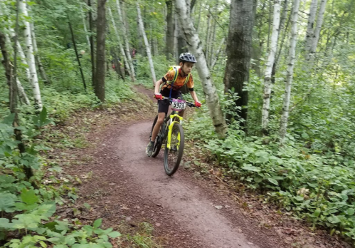 A young cyclist rides a mountain bike along a winding trail in a lush, green forest.