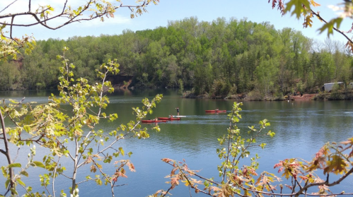 A serene lake surrounded by greenery, with two kayakers paddling in the calm water under a clear blue sky.