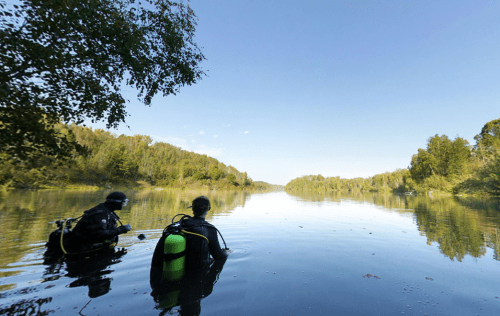 Two divers in wetsuits enter a calm lake surrounded by lush greenery under a clear blue sky.