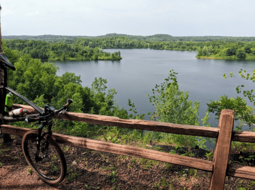 A bike rests against a wooden fence overlooking a serene lake surrounded by lush greenery and distant hills.