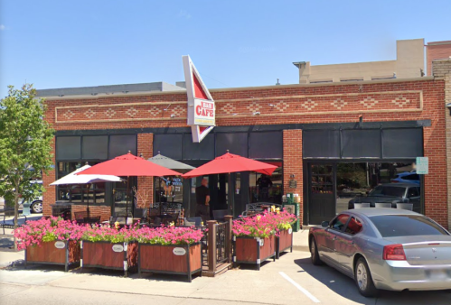 A brick restaurant with a large sign, outdoor seating under red umbrellas, and colorful flower planters.