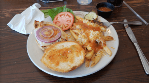 A plate with a grilled chicken sandwich topped with onion, a side of cheesy fries, lettuce, tomato, and a small cup of sauce.