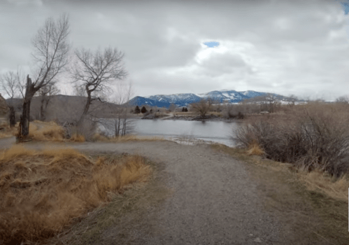 A gravel path leads to a calm river, surrounded by bare trees and mountains under a cloudy sky.