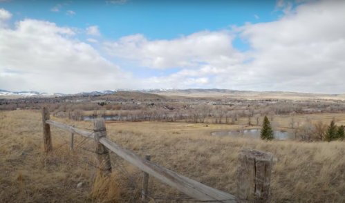 A scenic view of rolling hills, a river, and a wooden fence under a partly cloudy sky.