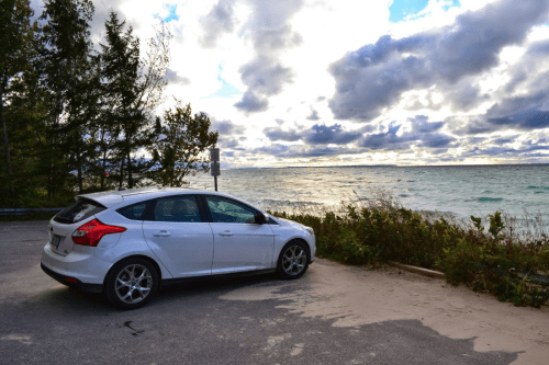 A white car parked near a shoreline with trees and a cloudy sky in the background.