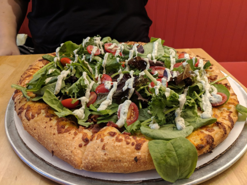A large pizza topped with fresh greens, cherry tomatoes, and drizzled with dressing on a cheesy crust.