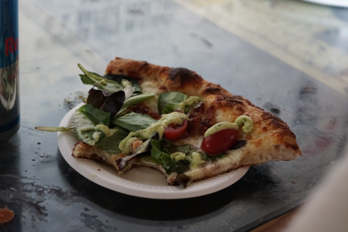 A slice of pizza topped with greens and cherry tomatoes on a paper plate, resting on a table.